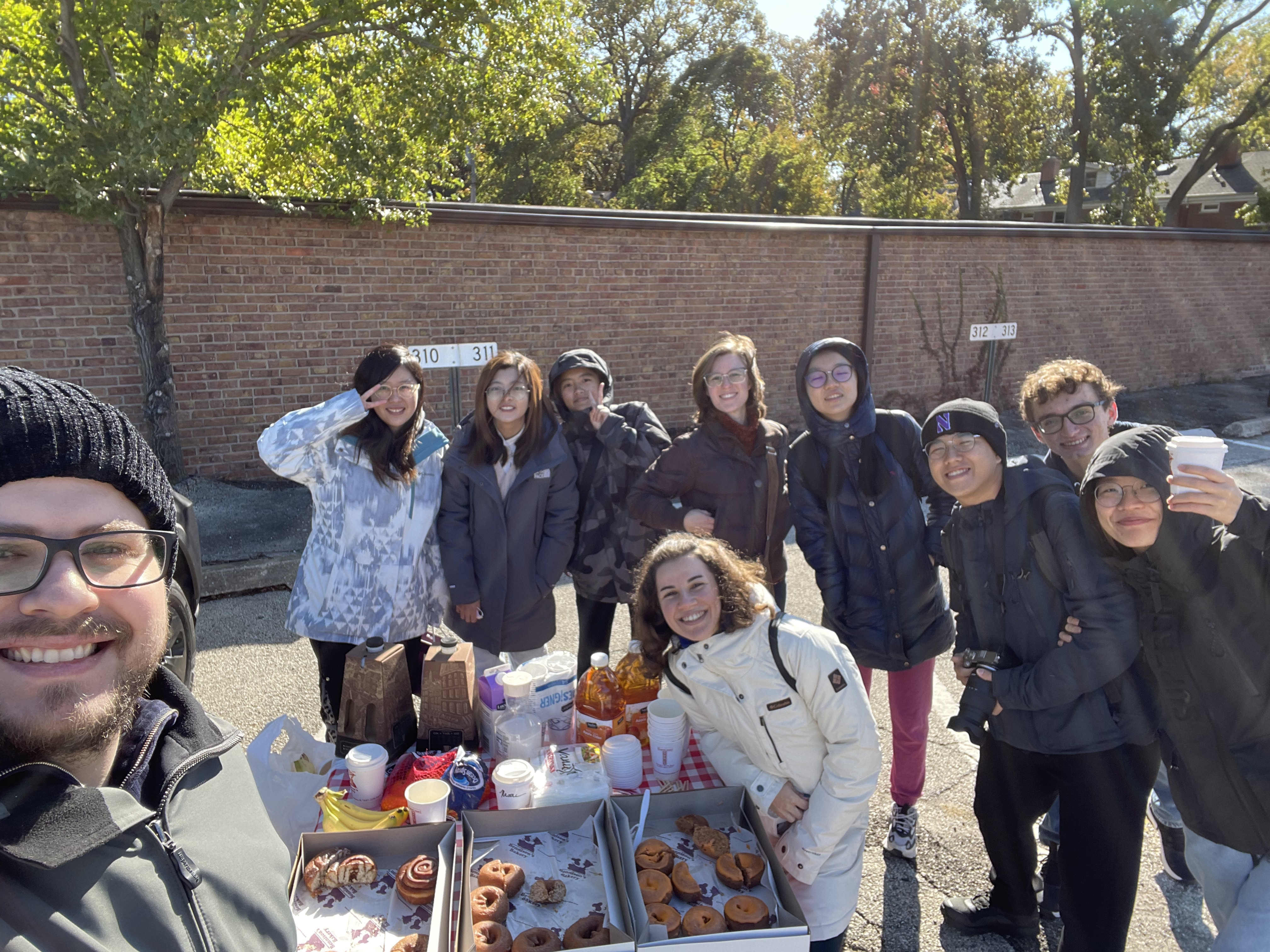 A group of PhD students in front of a table of pastries outside, smiling at the camera.